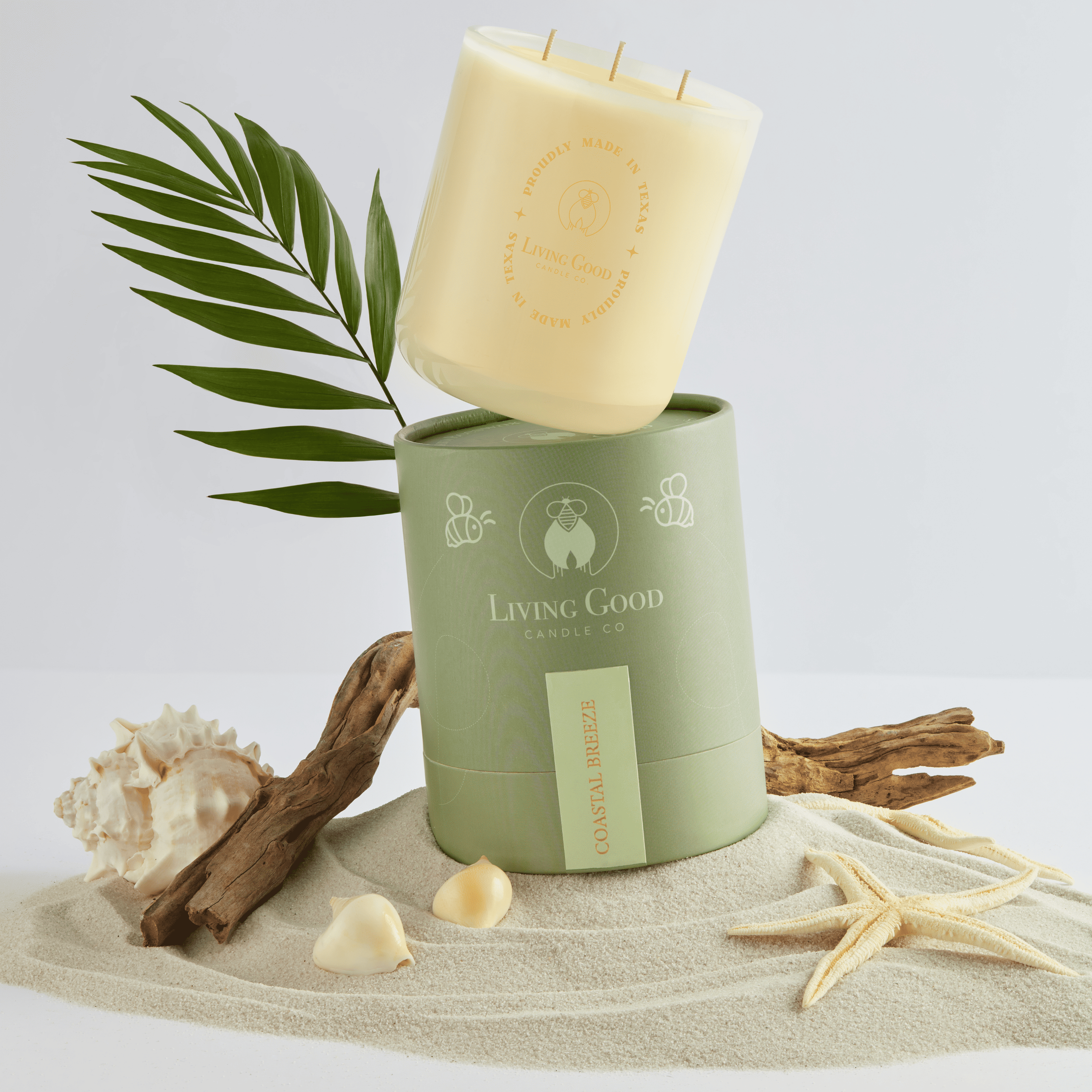 Living Good Candle Co. Coastal Breeze beeswax candle with beach-inspired packaging, displayed on sand with seashells, driftwood, and tropical leaves.
