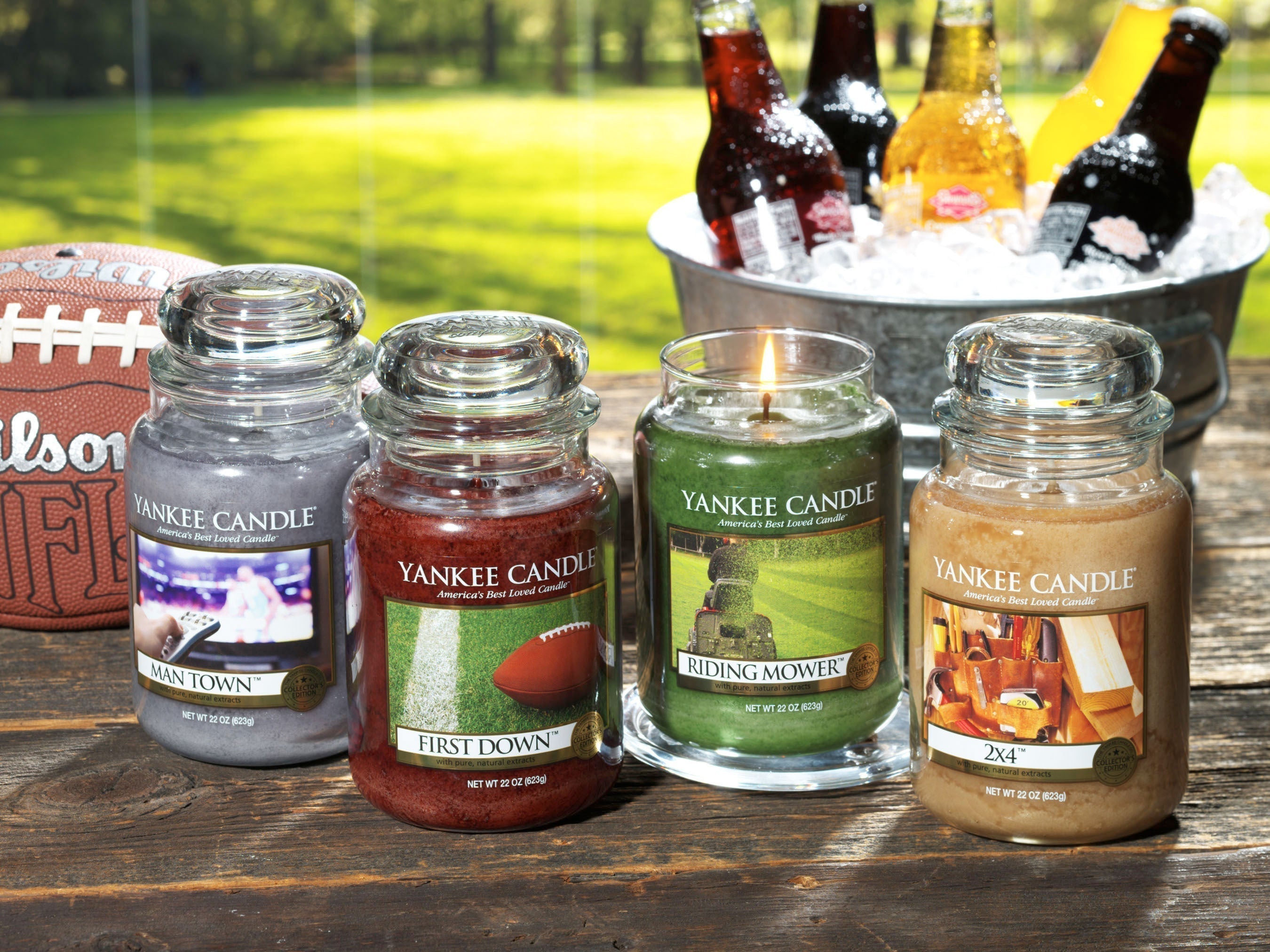 Four Yankee Candle jars arranged on an outdoor wooden table with a sunny backyard view, football, and a bucket of iced drinks in the background. Scents featured include Man Town, First Down, Riding Mower.