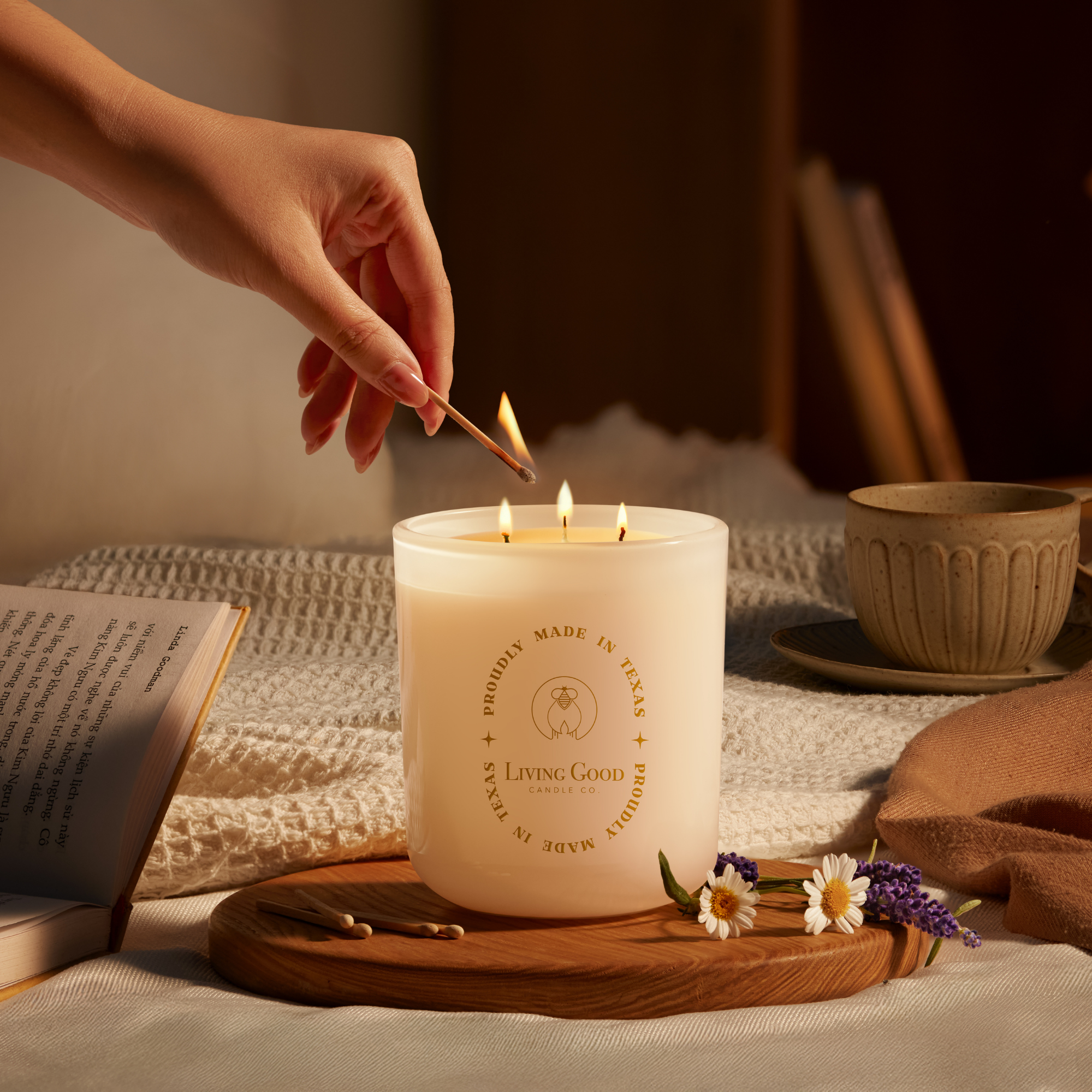 A hand lighting a three-wick white beeswax candle from Living Good Candle Co., surrounded by calming elements like a ceramic tea cup, chamomile flowers, lavender, and an open book—capturing a peaceful, toxin-free moment at home.