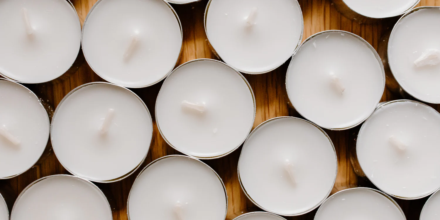 Flat lay of multiple paraffin tealight candles in metal tins, tightly arranged on a wooden surface, showcasing their smooth, white wax appearance.