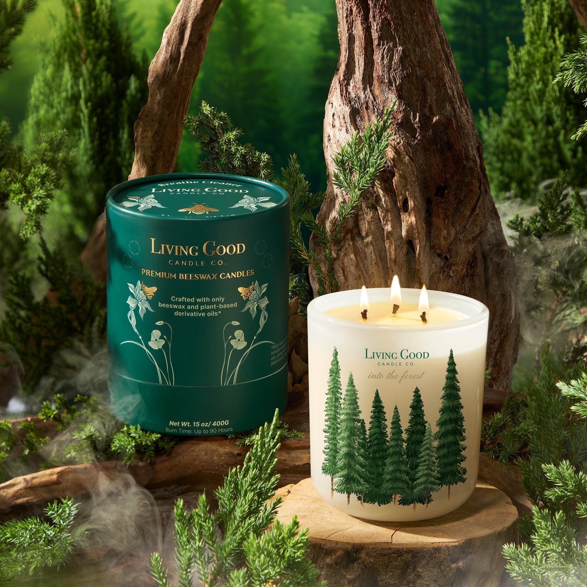 Living Good Candle Co. 'Into the Forest' beeswax candle with illustrated evergreen tree design, shown burning next to its green packaging tube in a lush forest-inspired setting with natural wood and greenery.