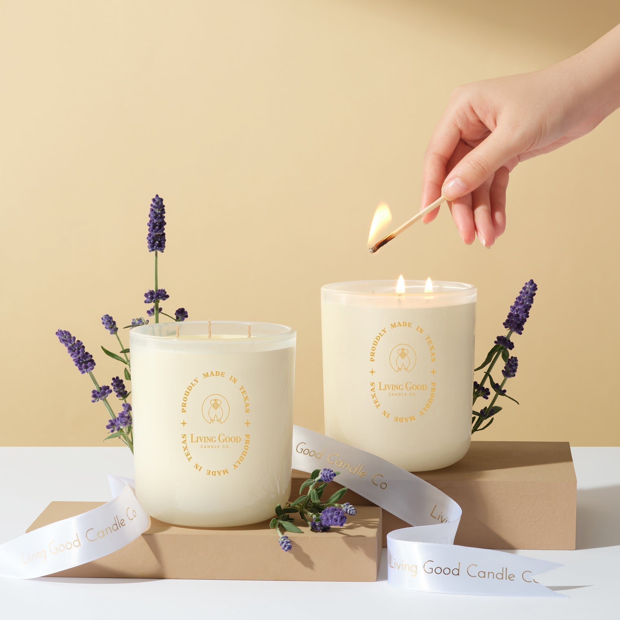 A hand lighting a white beeswax candle from Living Good Candle Co. next to another lit candle, both surrounded by lavender sprigs and branded ribbon, placed on beige platforms against a warm neutral background.