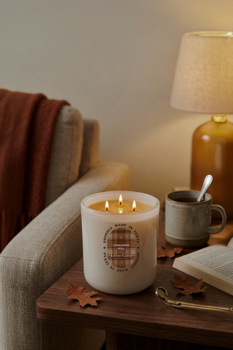Living Good Candle Co. 15 oz Cinnamon Roll three-wick beeswax candle glowing beside a book and coffee in a cozy fall home setting.