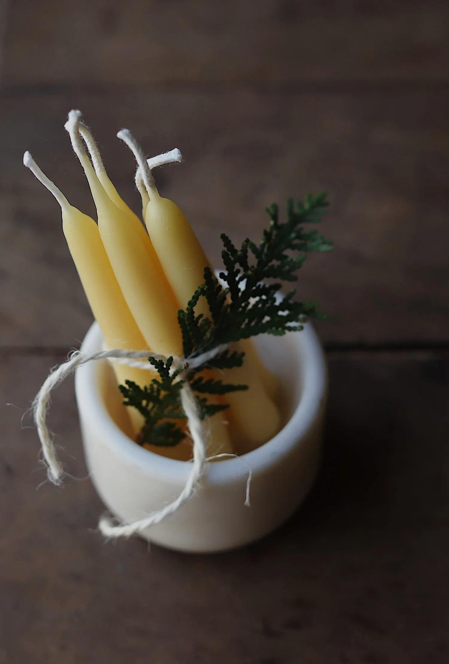A bundle of hand-dipped beeswax taper candles tied with twine and decorated with a sprig of greenery, displayed in a small white ceramic holder on a rustic wooden surface.