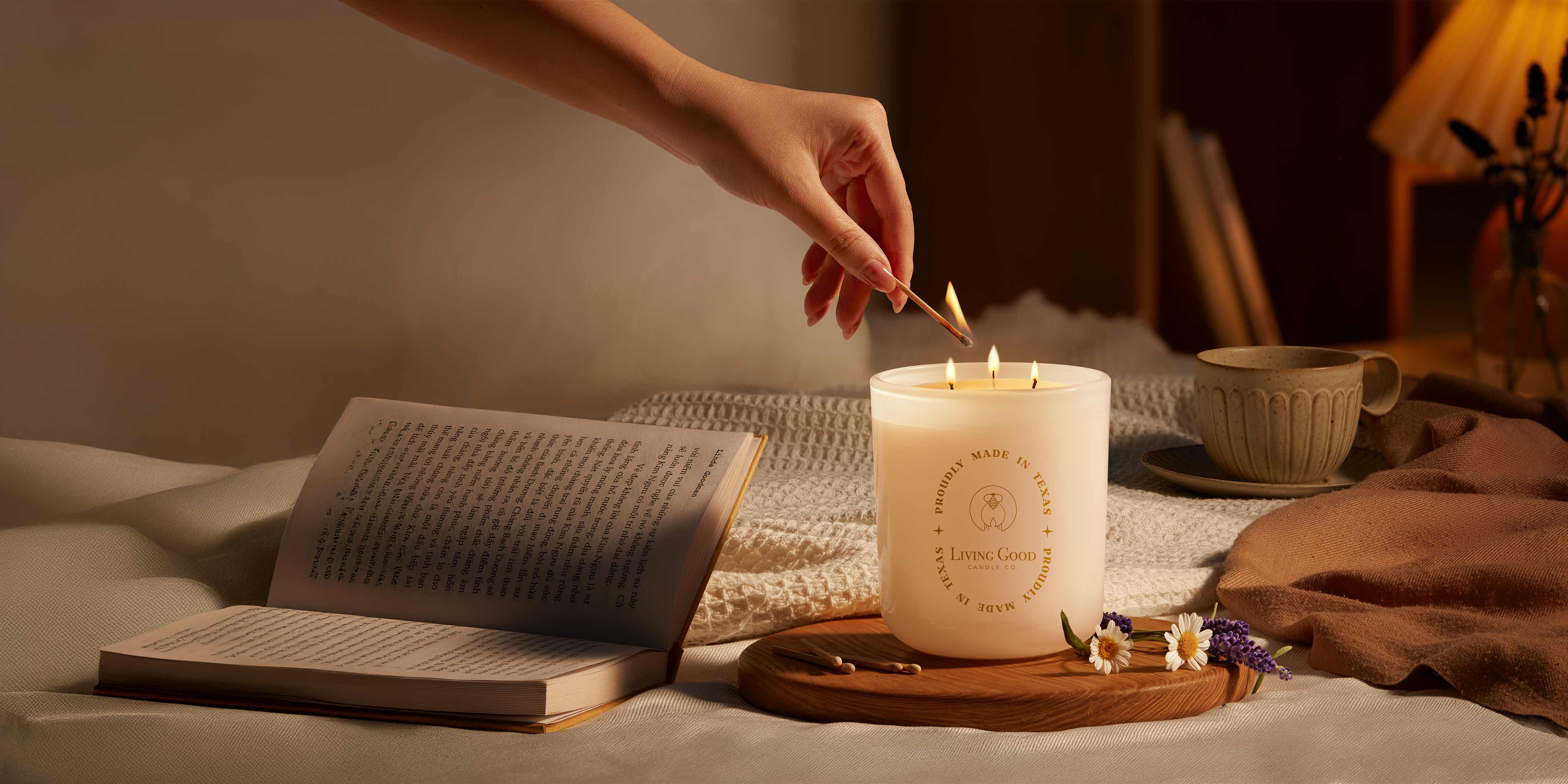 A cozy bedroom scene featuring a hand lighting a triple-wick beeswax candle from Living Good Candle Co., surrounded by an open book, a cup of tea, and soft ambient lighting.